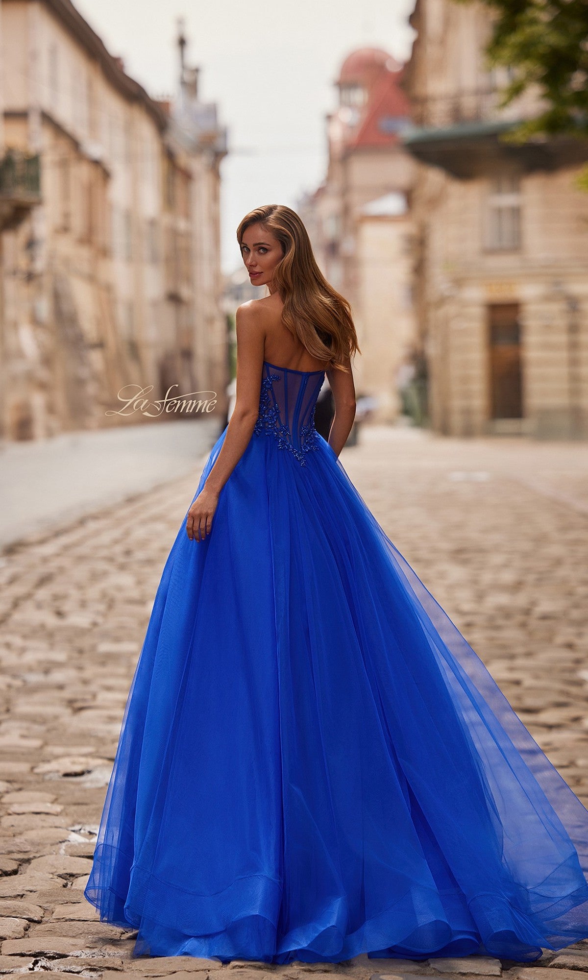 Wearing the La Femme 33363 long prom dress, a woman in a strapless ball gown with an A-line tulle skirt stands on a cobblestone street, glancing over her shoulder with historic buildings behind her.